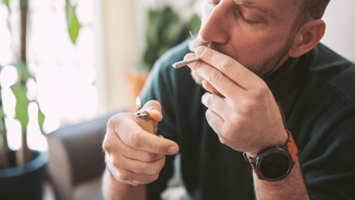 A man sits on his couch in a sunlit room and lights a legal cannabis joint