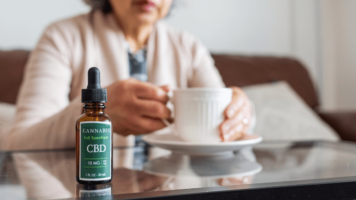 a CBD tincture bottle sits on a table in front of a senior woman holding a cup of tea